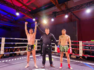 3 Personen in einem Boxring, der Kampfrichter in der Mitte, links und rechts ein Kämpfer, der Kämpfer links streckt als Sieger die Arme in die Luft