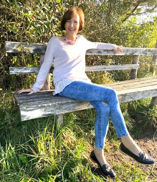 Margit Königsecker, Coach & Trainerin -  sitzt im Freien auf einer Holzbank und blickt in die Kamera. Trägt weißes Oberteil und Jeans. Im Hintergrund sind Bäume und Sträucher, die Sonne scheint