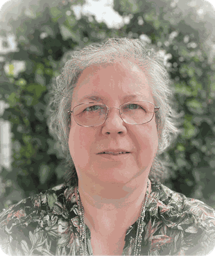 Porträt einer älteren Frau mit Brille und grauem, gewelltem Haar vor einer grünen, leicht verschwommenen Naturkulisse. Sie schaut ruhig und freundlich in die Kamera.