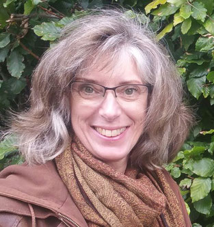 Joke Zonneveld, een stralende vrouw, lachende mond, met vol, bruin, tikje grijzend haar en groene ogen, bril, bruinejas en sjaal voor een groene beukenheg