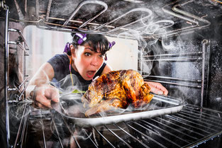 Frau mit Lockenwicklern in den Haaren zieht entsetzt ein verbranntes Brathähnchen aus dem Ofen. 