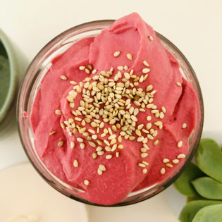 Pink Hummus. Pinker Rote Beete Hummus im Glas. Fotografiert von oben. Getippt mit beigen Sesamsamen. Im Hintergrund ist der Ansatz einer Pflanze zu sehen.