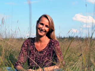 Engelcoach Carola Gaubatz im Schneidersitz auf einer Wiese mit hohen Gräsern ein blauer Himmel im Hintergrund