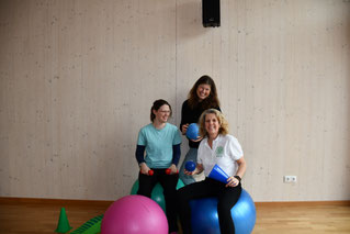 Drei Frauen sizten stehen mit verschiedenen Sportgeräten und schauen einladend fröhlich.