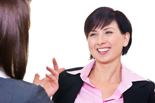 Eine Frau mit strahlendem Lachen, die von einer ihr gegenübersitzenden Frau etwas gesagt bekommt.