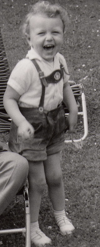 Peter Bach jr. in bayerischer Tracht im Alter von 4 Jahren: Ein kleiner Junge mit kurzen Lederhosen lacht mit Lockenkopf in einem schwarz/weißen Bild zur Kamera. Er steht auf einer Wiese vor einem Liegestuhl.