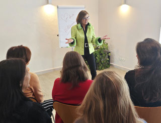 Margit Königsecker, Coach und Trainerin steht vor einer Gruppe von Frauen in einem Seminarraum - vor einem Flipchart und spricht zu den Teilnehmerinnen. Sie trägt schwarze Hose und schwarzes Oberteil und darüber einen grünlich/selben Blazer