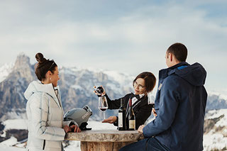Il sommelier sulle piste dell'Alta Badia serve il vino a due appassionati di sport invernali; degustazione all'aperto con panorama sulle Dolomiti e cime innevate.