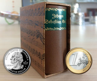 A tiny biography of Bach is visible at an angle from the front. It is in a slipcase. At the front right and left of it, a euro coin and a US quarter coin indicate its size.