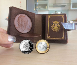 A tiny Bach mini-book is visible in an open leather ensemble. On the left is a clay medal with Bach's portrait, on the right is the small book with the Bach Seal on the cover. At the front are two coins for a size comparison: € 1 and 1 US quarter.