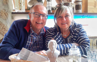 Peter on the left and Renate Bach on the right are smiling to the camera. They are both sitting at a table. There is a small Bach bust in the foreground. Both wear mostly blue clothes.