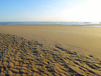 Ein breiter rot brauner Sandstrand zeichnet die Wellen des Wasser nach. Am Horizont leuchtet das Meer tiefblau unter der weißen Sonne