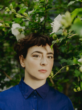 Die Autorin Kate Zambreno trägt eine Kurzhaarfrisur und eine hochgeschlossene blaue Bluse. Sie steht vor einem Rosenbusch.