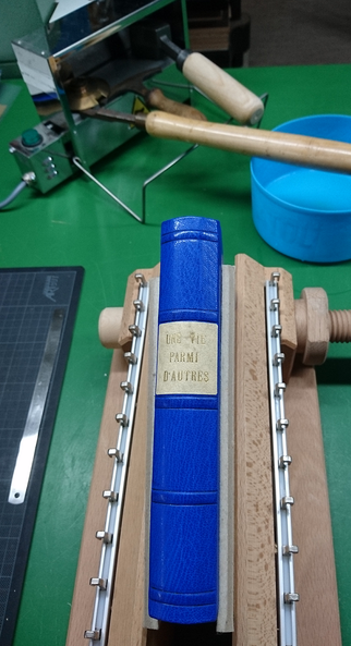 zu sehen ist ein blaues Lederbuch, eingespannt in eine Klotzpresse. Davor sind Vergoldezwerkzeuge auf einem Ofen zu sehen. Diese werden darin heiß gemacht. Es folgt eine Handvergoldung in Blind.