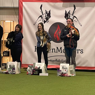 Drei Personen stehen mit Hunden vor einem Banner mit dem Schriftzug 'HundenMoers' und einem Flammensymbol, vor ihnen liegen Preise und Futterbeutel auf dem Boden.