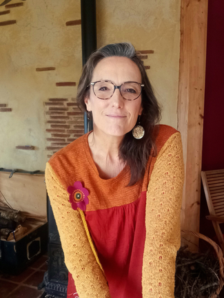 Portrait de Laurence de face, regard profond, accueillant et sourire doux, elle pose devant un mur de terre paille, enduit terre et briquettes, ambiance naturelle, douce et chaleureuse.