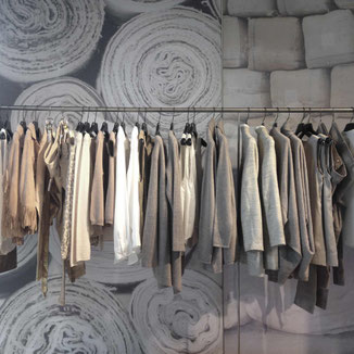 wunderschön assortierte Kleider in weiss, beige , braun hängen an einer Kleiderstange aus CNS. Im Hintergrund ist ein Spanntextil mit farblich passenden natürlichen Stoffrollen und Rohbaumwolle zu sehen. 