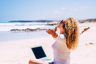 Frau sitzt mit Laptop am Strand. Workation. Arbeiten im Ausland. Mit Langzeit-Reiseversicherung und Krankenversicherung für lange Auslandsreisen.