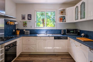 Cuisine spacieuse et lumineuse avec des meubles blancs classiques, un plan de travail bleu et une grande fenêtre offrant une vue sur la verdure. L'aménagement fonctionnel et l'éclairage naturel sont mis en valeur pour la photographie immobilière.