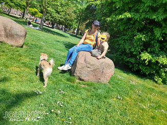 Eine Frau mit Kind sitzt auf einem Stein in einem Park. Davor steht ein kleiner, blonder Hund.
