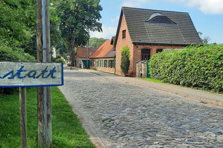 Eine Dorfstraße ist mit grobem Kopfsteinpflaster gepflastert. Am Rand stehen gepflegte Häuser mit roten Backsteinmauern, grünen Fensterläden und grünen Hecken. 