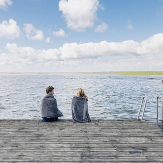 Zwei in Handtücher gehüllte Personen sitzen auf einem Holzsteg an der Nordsees, im Hintergrund grüne Deiche und Küstenlandschaft