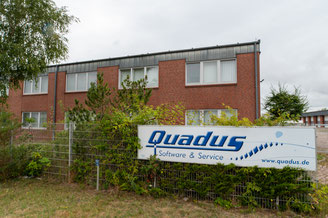 Zweistöckiges Bürogebäude aus rotem Backstein mit einem Schild 'Quadus Software & Service www.quadus.de' an einem Metallzaun im Vordergrund