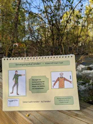 Der gelbe Kalender steht auf einem Hopygeländer einer Brücke. Im Hintergrund ein Bach und Bäume. Auf dem Blatt zwei Bilder von einer Frau und einem Mann die eine Übunge machen. Text: Bewegungskalender Koordination.