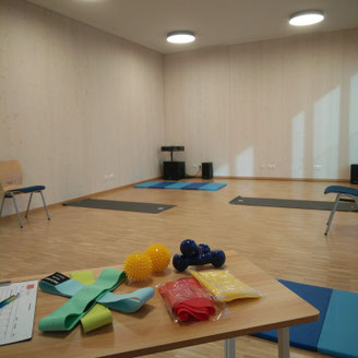Blick in den Trainingsraum mit verschiedenen kleinen Trainingsgeräten auf einem Tisch(Bänder und Bälle) und Matten auf dem Boden, sowei Stühle.