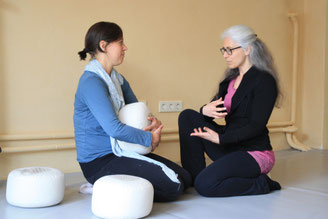 Eine Frau zeigt einer anderen etwas, eine Frau hält ein Yogakissen, beide sitzen auf dem Boden in einem Yogastudio oder Coachingstudio