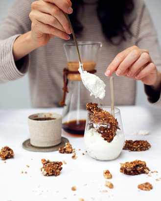 Someone sitting behind a Chemex coffee pot, with a handmade plate and cup, and a glass of yoghurt with granola. Pieces of granola are scattered across the table.