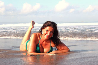 Im Bild ist eine dunkelhaarige Frau im Bikini. Sie liegt am nassen Sand am Strand - unmittelbar vor den heranlaufenden Wellen - mit angewinkeltem Bein