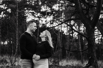 portret in het bos van zwanger koppel dat elkaar verliefd aankijkt, de wind speelt door de haren van de vrouw-zwangerschapsfotografie-©Fotografils-byIlseWagemakers-fotograaf-based in Essen, België