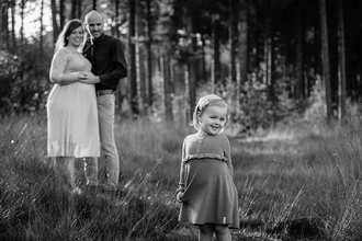 meisje kijkt guitig in de lens terwijl haar zwangere mama en papa trots vanop de achtergrond toekijken-zwangerschapsfotografie-©Fotografils-byIlseWagemakers-fotograaf-basedinEssen