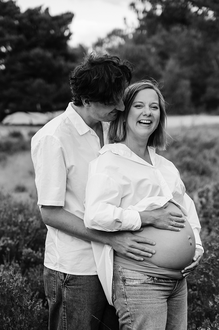 portret van zwanger koppel in de heide waarbij de man de vrouw iets in haar oor fluistert waardoor ze lacht-zwangerschapsfotografie-©Fotografils-byIlseWagemakers-fotograaf-based in Essen, België