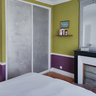 Chambre rénovée dans une maison du Val-d'Oise avec murs vert olive et soubassement prune, placard enduit, cheminée ancienne noire et miroir, projet d’architecture d’intérieur.