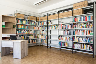 Ein Foto der Bibliothek. Zwei Regalwände mit Büchern. Ein Schreibtisch.