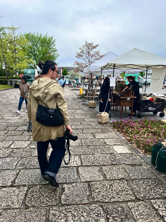 帆布バッグのCargo Shipのオリジナル帆布バッグCarryーOnのブラックを斜めにかけ、髪を括った男性がカメラを片手に山口アーツ&クラ仏２０２６のAブロックの石畳を歩く様子
