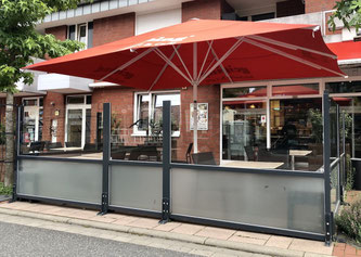 Klassischer Glas-Windschutz mit grauen Rahmenprofilen auf einer Restaurant-Terrasse unter einem roten Sonnenschirm