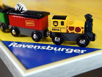 Eine Holzeisenbahn steht vor dem dreieckigen Logo des Spieleherstellers Ravensburger. Foto: Felix Kästle/Illustration