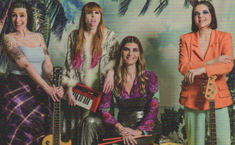 Die vier Bandmitgliederinnen mit Instrumenten und schrillen Klamotten vor einer Palmenfototapete.