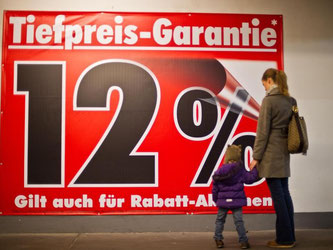 Eine Frau betrachtet mit ihrer Tochter ein Werbebanner mit «Tiefpreis-Garantie». Foto: Jan-Philipp Strobel/Archiv/Symbol
