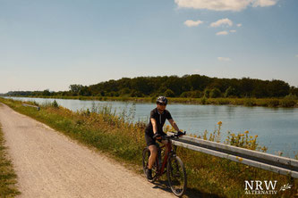 Frau auf einem Fahrrad, im Hintegrund Wasser und Bäume.