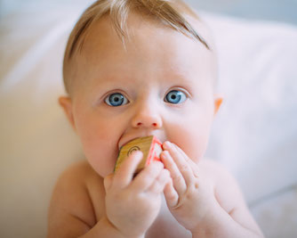 Ein Baby welches geradewegs in die Kamera schaut. Es hält mit beiden Händen einen Holzwürfel fest, den es mit dem Mund abtastet.
