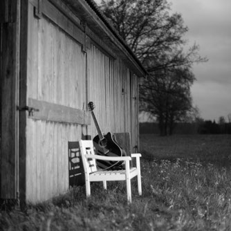 Eine Parkbank vor einer alten Holzhütte. Darauf eine Gitarre und eine Cajon.