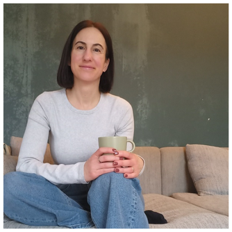 Michaela Dauscher sitzt auf dem Sofa, hat ein graues Shirt an und eine blaue Jeans. Ihre braunen Haare sind schulterlang, sie lächelt.