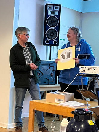Stefanie Eilers (rechts im Bild) überreicht Klaus Börgmann (links im Bild) einen Ausstellungskatalog und eine Grafik mit zwei feiernden Wildschweinen als Dank für seinen Vortrag