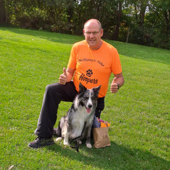 Mann in orangefarbenem T-Shirt mit Aufschrift 'Wolfgang & Mika Hoopers' kniet auf grünem Rasen neben schwarz-weißem Border-Collie-Hund mit herausgestreckter Zunge.
