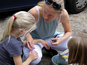 Eine Frau kniet, sie spricht zu zwei Kindern, in ihrer Hand ein Vogel