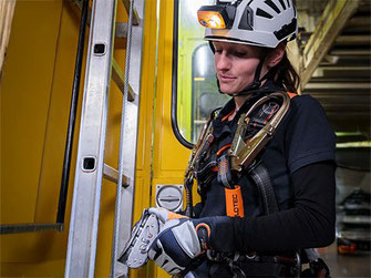 Eine Frau mit Helm, Stirnlampe, Auffang- und Haltegurte und Y-Verbindungsmittel mit Bandfalldämpfer arbeitet an einer Steigleiter in einem Hochregallager
